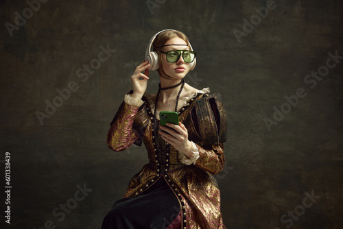 Fototapeta Portrait of young beautiful girl in vintage dress, trendy sunglasses, headphones and mobile phone against dark green background
