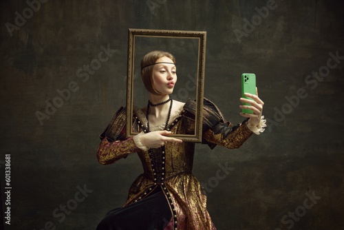 Portrait of pretty young girl, royal person in vintage dress holding picture frame and taking selfie with phone against dark green background. Concept of history, renaissance art, comparison of eras