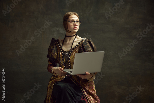 Portrait of young pretty girl, queen in vintage dress posing with laptop over dark green background. Education, freelance job, business. Concept of history, renaissance art remake, comparison of eras