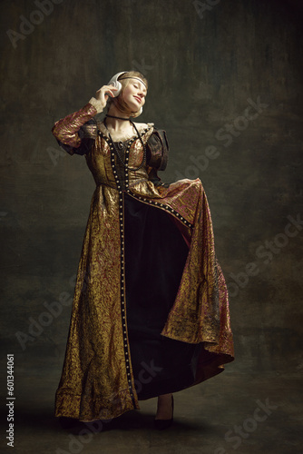 Full-length portrait of young girl, royal person, princess in vintage dress, listening to music in headphones over dark green background. Concept of history, renaissance art remake, comparison of eras