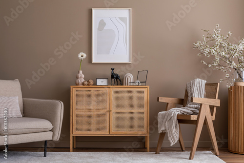 Fototapeta Naklejka Na Ścianę i Meble -  Warm and cozy living room interior with mock up poster frame, rattan sideboard, wooden armchair, beige sofa, bright carpet, plaid, cherry blossoms and personal accessories. Home decor. Template.