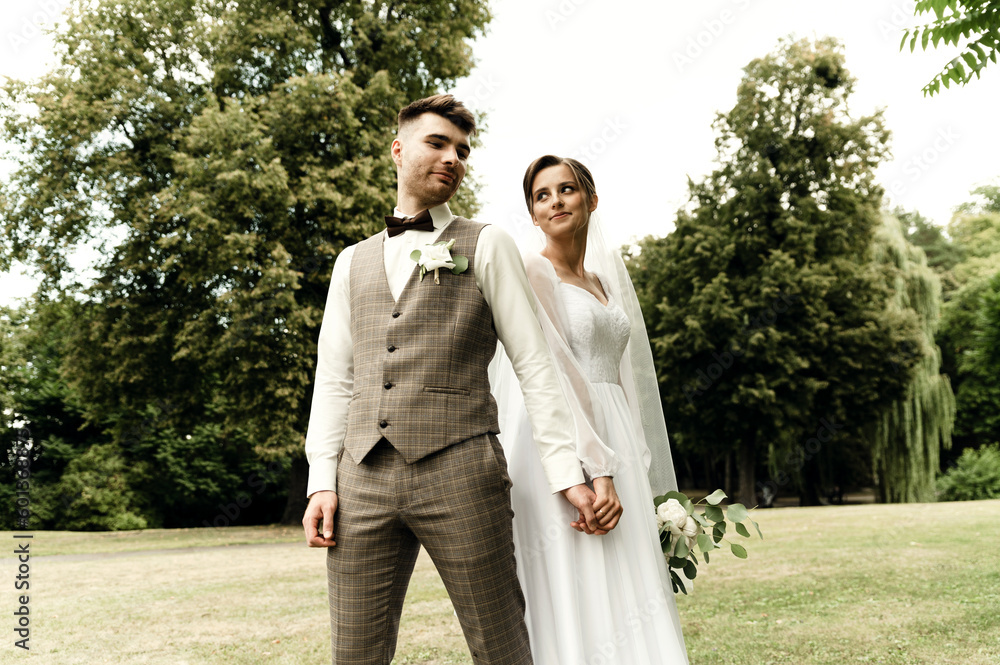 Fototapeta premium modern newlyweds in rustic style. wedding couple holding hands standing with their backs to each other. wedding outside in summer. attractive wedding couple