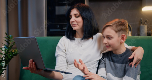 Beautiful woman with cute 9-year-old boy sitting together on couch holding laptop while talking on video chat with family. Happy family and modern technology photo