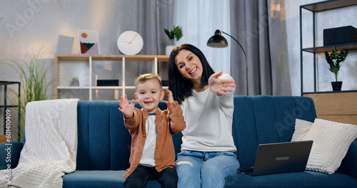 Adorable smiling happy mother and her little in high spirits son sitting together on the sofa at home and waving hands into camera to say hello photo