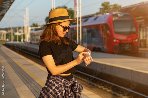Tourist woman wearing straw hat standing on railway platform waiting train and using her smart watch.