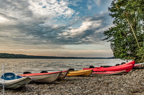 North Hatley, QC, Canada - September 5, 2014: Red, yellow, blue and green kayaks sit on a pebbly beach beside a stand of trees as the sun rises with dramatic clouds over Lake Massawippi photo