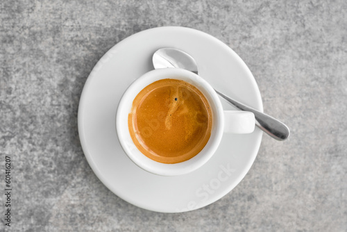 White cup with fragrant strong espresso coffee with thick foam in a traditional Greek coffee shop. Top view of coffee with saucer on gray marble table, idea for background or poster