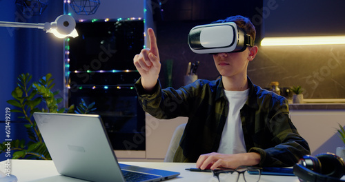 Teen blond caucasian boy in VR glasses sitting in evening kitchen and having VR headset. Male moving hands on virtual screen applying augmented reality goggles.