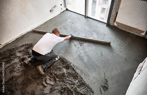 Top view of male construction worker placing screed rail on the floor covered with sand-cement mix. Man smoothing and leveling surface with straight edge while screeding floor in apartment. photo