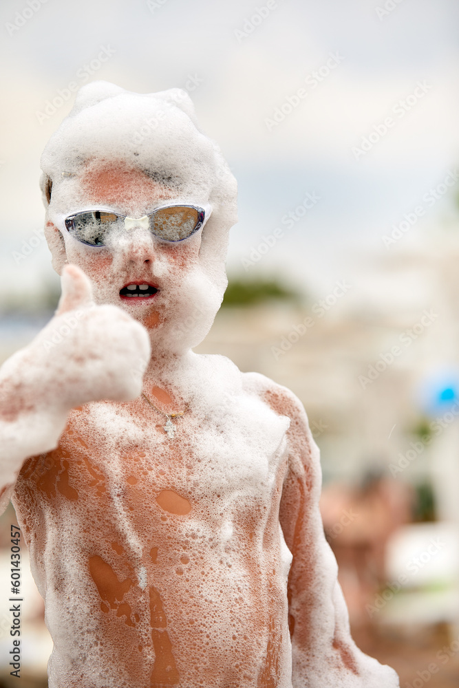 Happy little boy with goggles and foam on his head having fun in a water park, in wet swimwear at a foam party or holiday on a sunny hot summer day - obrazy, fototapety, plakaty 