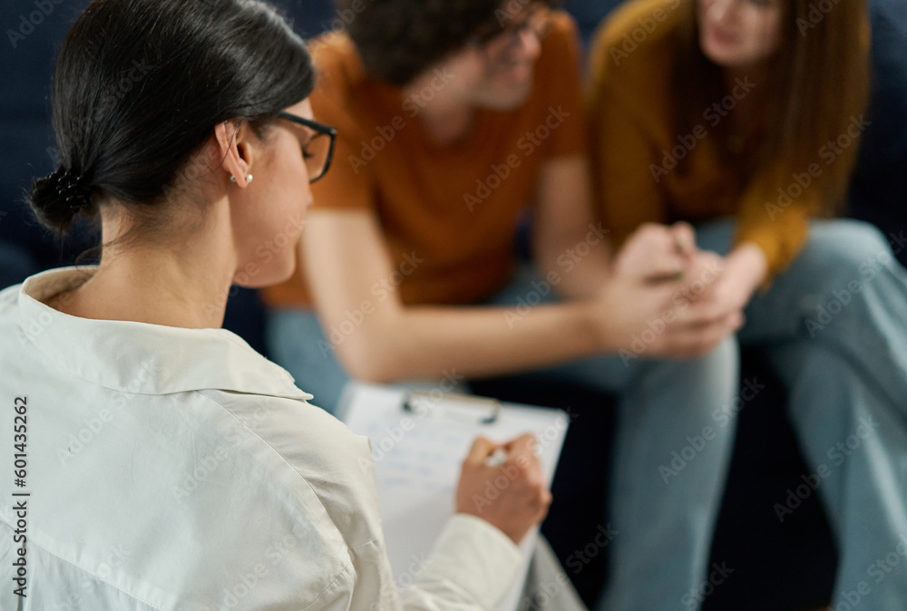 Rear view of female psychologist having counseling or therapy session with a clients. Woman listening and takes notes during couple speaks. Selective Focus Rear view of female psychologist having counseling or therapy session with a clients. Woman listening and takes notes during couple speaks. Selective Focus