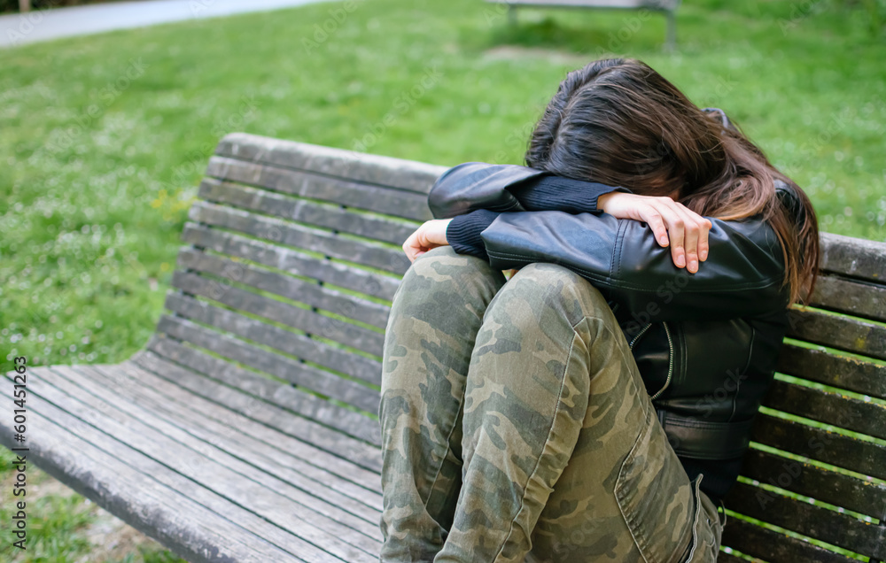 Young woman sad and depressed situated on park bench with arms and head on knees Young woman sad and depressed situated on park bench with arms and head on knees