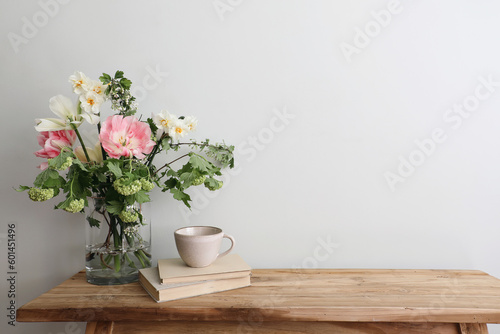 Moody spring still life. Wooden bench, table composition with cup of coffee, tea and old books. Beautiful floral bouquet with white, pink tulips, daffodils. Hawthorn, green guelder rose flowers. Wall.