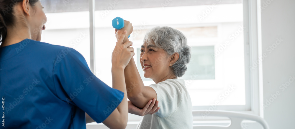 Fototapeta premium Young physiotherapist helping senior mature asian woman grey hair work out with dumbbells, to recover from injury at health centre in physical therapy session.