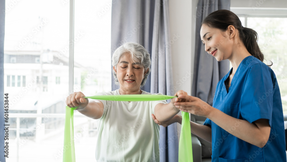 Obraz premium Young physical therapist caregiver assisting mature asian woman grey hair doing exercise with elastic bands at physiotherapy clinic.