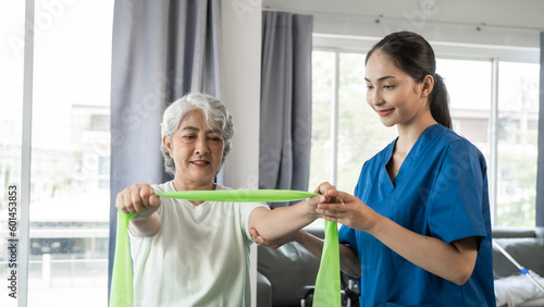 Young physical therapist caregiver assisting mature asian woman grey hair doing exercise with elastic bands at physiotherapy clinic.