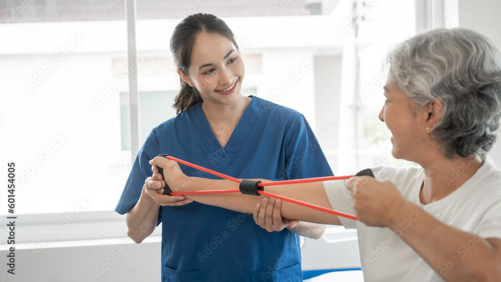 Young physical therapist caregiver assisting mature asian woman grey hair doing exercise with elastic bands at physiotherapy clinic. Young physical therapist caregiver assisting mature asian woman grey hair doing exercise with elastic bands at physiotherapy clinic.
