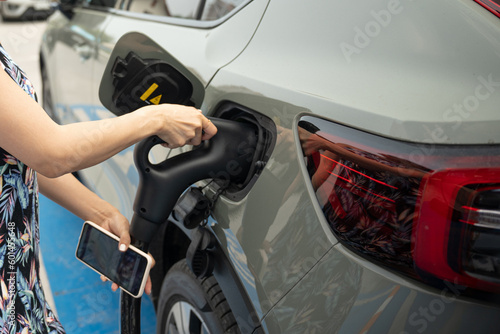 Selective focus of a woman's hands with a cell phone, holding a power cable supply plugged into a blurred green metallic car's charging port that parked at a charging station to recharge electricity.