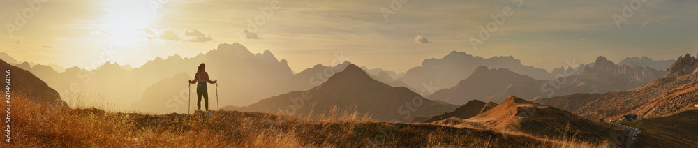 Panorama of  sunset mountains and a woman standing with trekking poles.