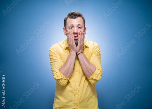 Awakening, man waking up and holding hands on cheeks over blue background, dresses in yellow shirt