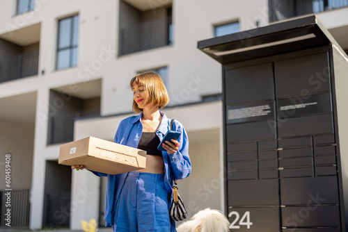 Young woman using smart phone while standing with a parcel delivered with post office machine with automatic lockers. New technologies in delivery service, self picking photo