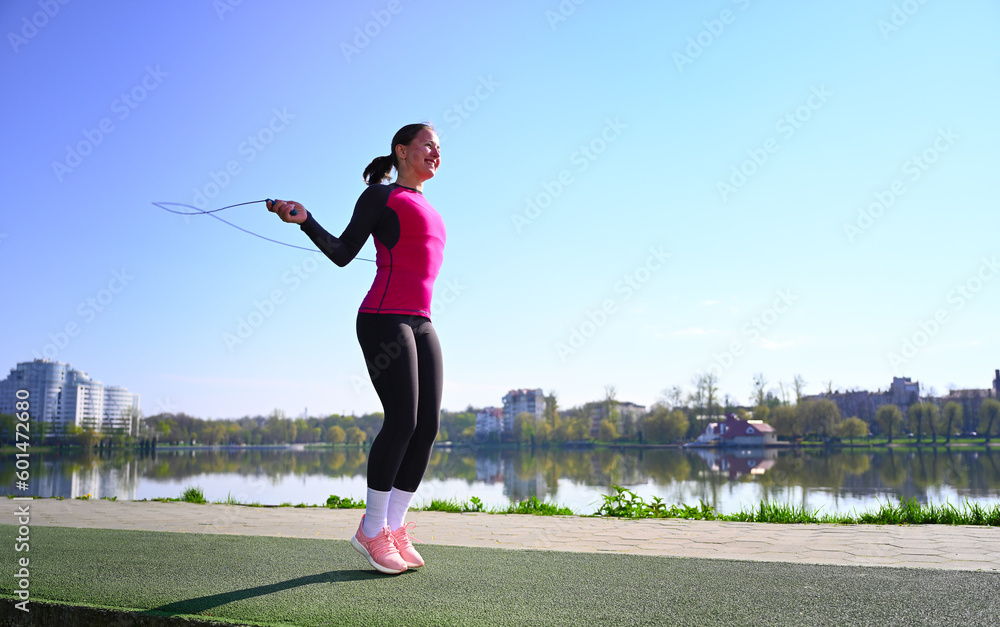 A young woman jumps on a skipping rope near the serene lake, with a focused expression on her face. Her active wear suggests her dedication to fitness and her determination to achieve her goals
