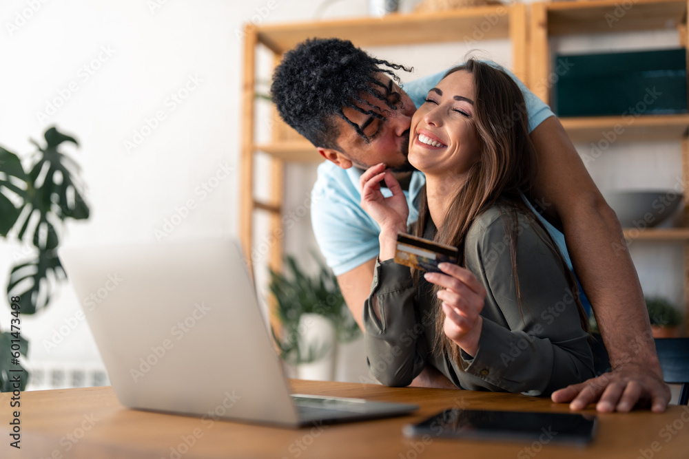 Smiling attractive young woman enjoying love kisses and company of her affectionate handsome husband while doing online shopping on laptop at home. Couple satisfied with online shopping.