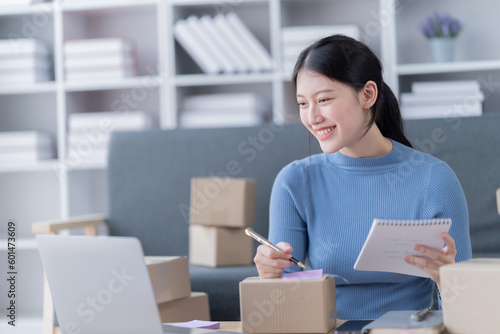 Asian business woman siting on sofa using a laptop and notebook checking order online shipping boxes at home. Starting SME Small business entrepreneur freelance. Online business, SME Work © Tj