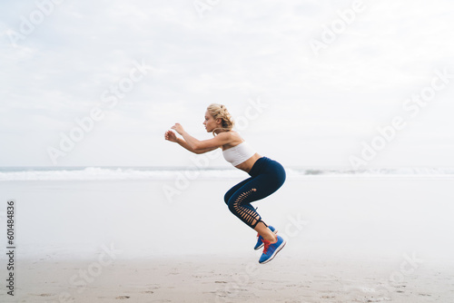 Blonde female athlete in tracksuit doing cardio exercises keeping body shape in tonus, determined sportswoman jumping spending time at seashore for training muscles strength and losing weight