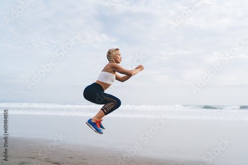Caucasian female athlete in tracksuit doing cardio exercises keeping body shape in tonus, young sportswoman jumping spending time at seashore for training muscles strength and losing weight