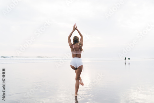 Back view of slim female in sportswear enjoying free time for training body vitality and flexibility during morning at coastline beach, blonde fit girl stretching muscles during yoga practice