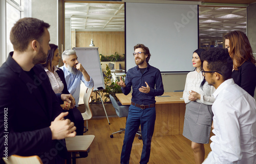 Young man business coach trainer talking to group of people in office standing around him and listening. Office workers, entrepreneurs on corporative training, team building seminar or master class. photo