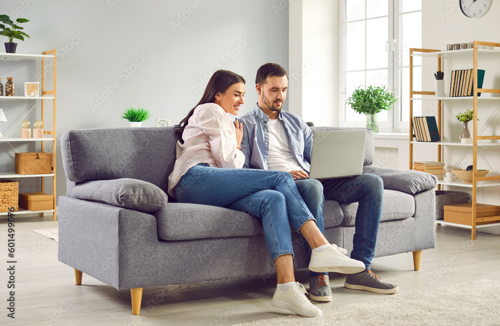 Happy family couple using laptop computer sitting on couch at home. Smiling young husband and wife looking at laptop screen while having video call, shopping online, browsing web, watching movie Happy family couple using laptop computer sitting on couch at home. Smiling young husband and wife looking at laptop screen while having video call, shopping online, browsing web, watching movie