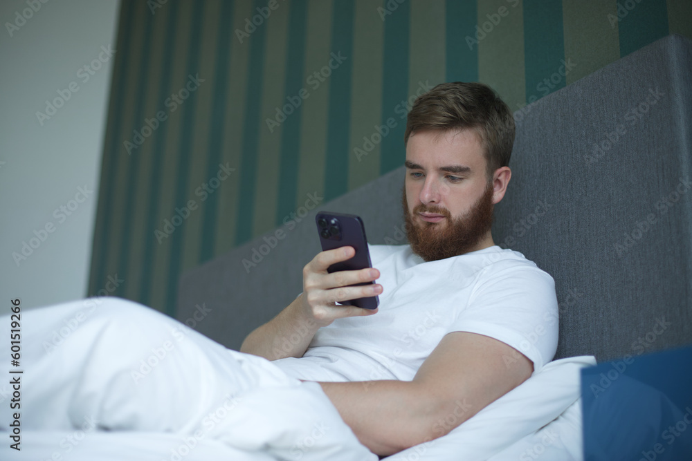 Young bearded man is using cell mobile phone at home in bed in bedroom, looking at screen of smartphone Young bearded man is using cell mobile phone at home in bed in bedroom, looking at screen of smartphone