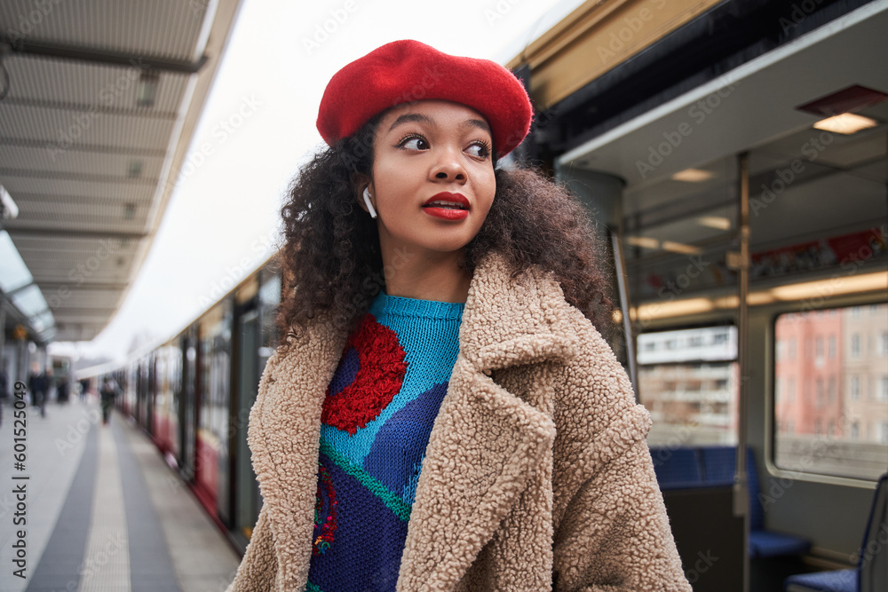 Young woman with curly hair wearing red hat standing at the tram station and listening music Young woman with curly hair wearing red hat standing at the tram station and listening music