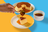 One hand holds a spoon with caramel syrup over a pudding dish, pour caramel syrup over the pudding, a cup of caramel on a yellow and blue background