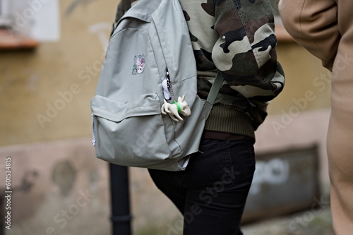 Zbliżenie na osobę niosącą szary plecak z zawieszką w kształcie psa, ubraną w kurtkę moro na tle miejskiej ulicy