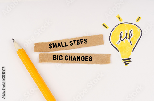 On a white surface, a luminous light bulb and pieces of paper with the inscription - small steps big changes