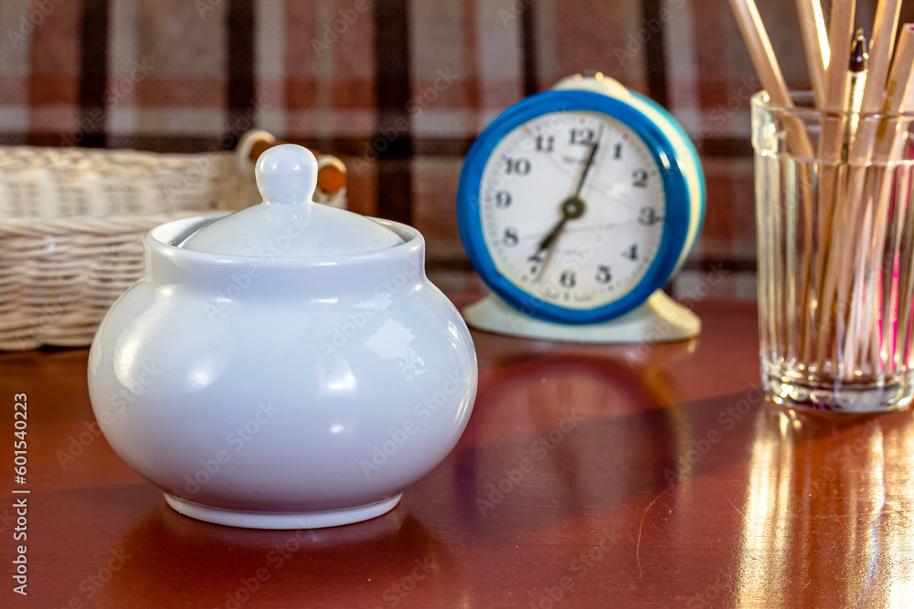 A sugar bowl, an old mechanical alarm clock, a glass with pencils are on the table