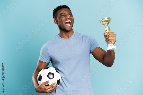 Portrait of overjoyed African American man, soccer player holding ball and trophy cup, celebration success isolated on blue background. Sport victory, winning competition concept photo