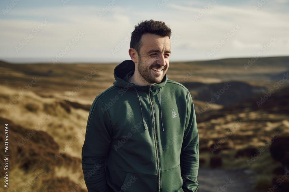 Handsome bearded man in green jacket and hood standing on top of a mountain and smiling