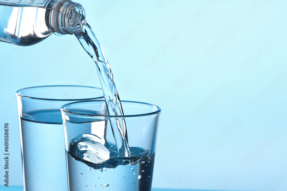 Pouring water from bottle into glass on light blue background, closeup. Space for text