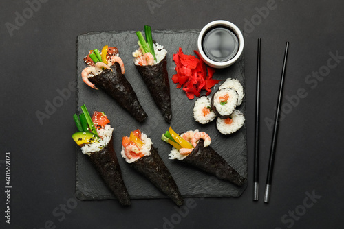 Slate board with tasty sushi cones, rolls, soy sauce, ginger and chopsticks on dark background