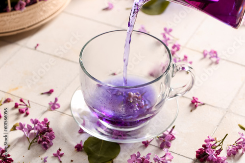 Pouring of hot lilac tea into glass cup and flowers on light tile table  closeup