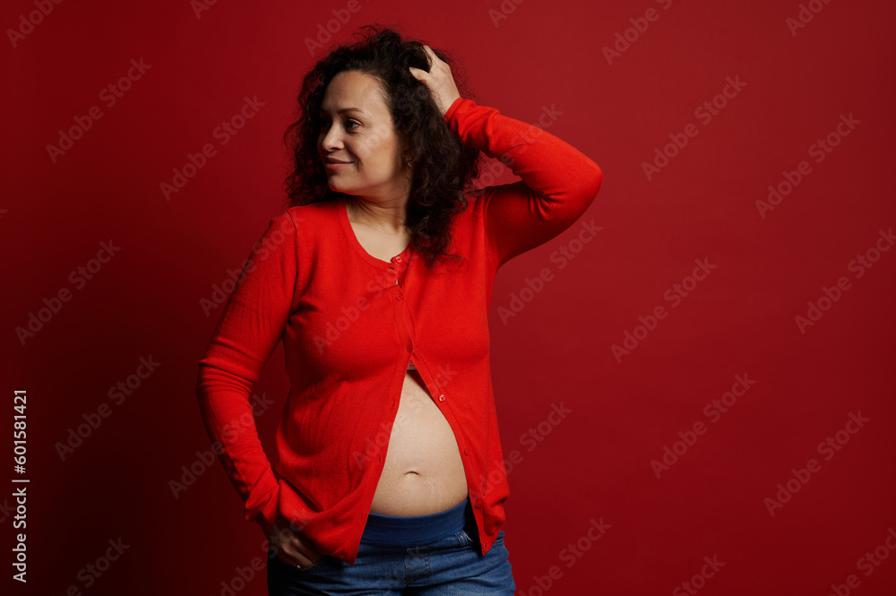 Confident portrait of a multi ethnic mature pregnant woman in red shirt and blue casual jeans, smiling looking aside, posing bare belly on isolated red background. Pregnancy fashion. Maternity concept Confident portrait of a multi ethnic mature pregnant woman in red shirt and blue casual jeans, smiling looking aside, posing bare belly on isolated red background. Pregnancy fashion. Maternity concept