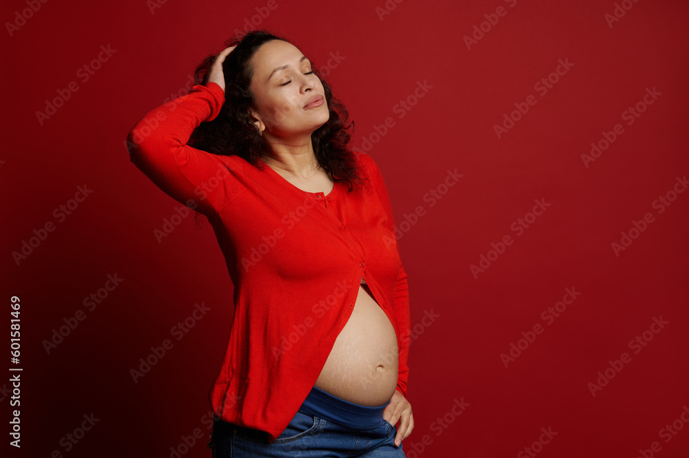 Fashion portrait of multi ethnic beautiful pregnant woman, expectant mother posing with closed eyes and bare belly over red isolated background. Happy Mothers Day. Pregnancy. International Women's Day Fashion portrait of multi ethnic beautiful pregnant woman, expectant mother posing with closed eyes and bare belly over red isolated background. Happy Mothers Day. Pregnancy. International Women's Day