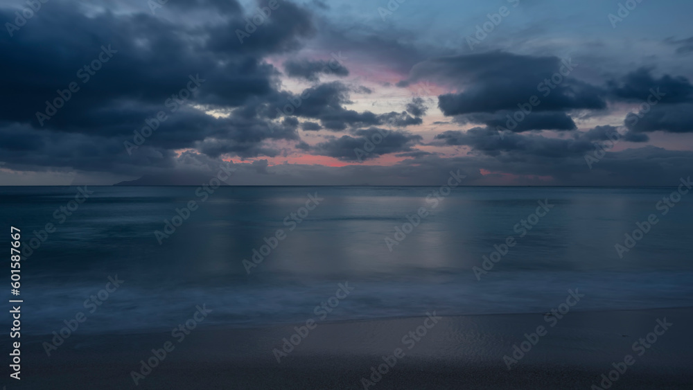Evening twilight on a tropical beach. Foam of waves on the sand. The silhouette of the island on the horizon. The sky is highlighted pink. Shades of blue. Seychelles. Mahe. Beau Vallon