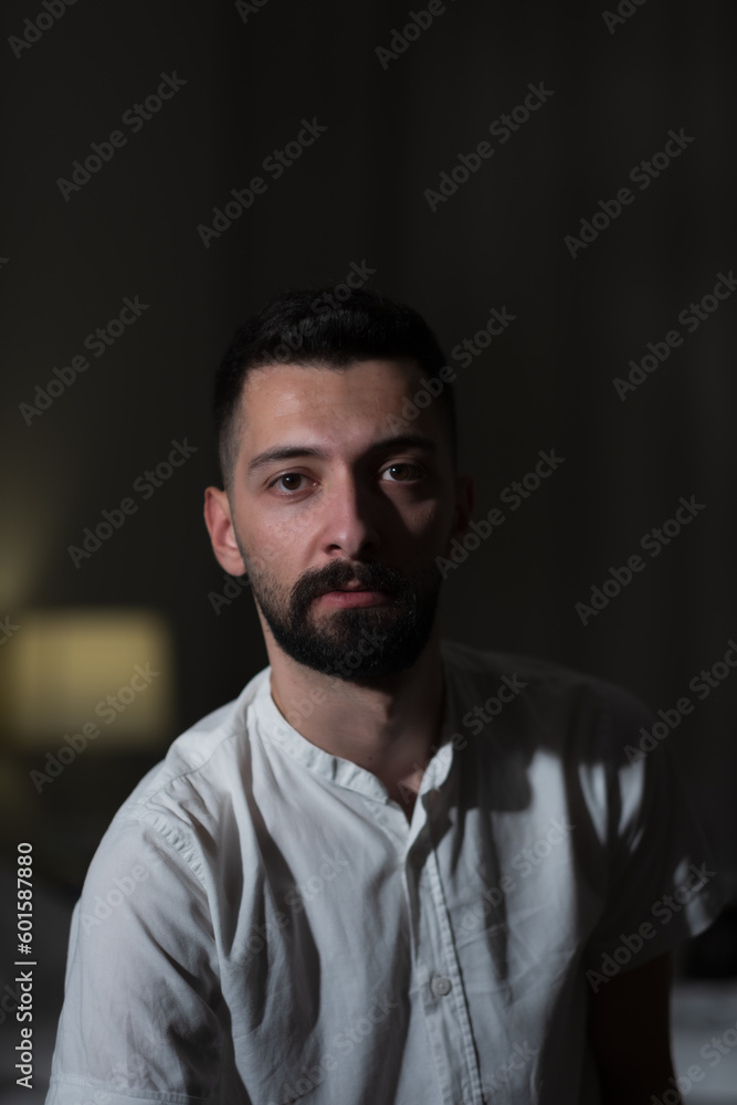 A young man from Syria sits indoors, and leans forward to pose for the camera. The background is dimly lit, but a bedside lamp is visible. His dark eyes and black beard are dramatized by the lighting. A young man from Syria sits indoors, and leans forward to pose for the camera. The background is dimly lit, but a bedside lamp is visible. His dark eyes and black beard are dramatized by the lighting.