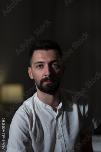 A young man from Syria sits indoors, and leans forward to pose for the camera. The background is dimly lit, but a bedside lamp is visible. His dark eyes and black beard are dramatized by the lighting.