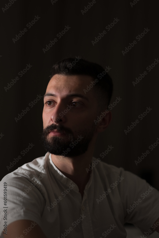 Looking off into the distance with a contemplative, melancholy expression, a young Syrian man poses in a dimly lit room with a single light source illuminating his face.